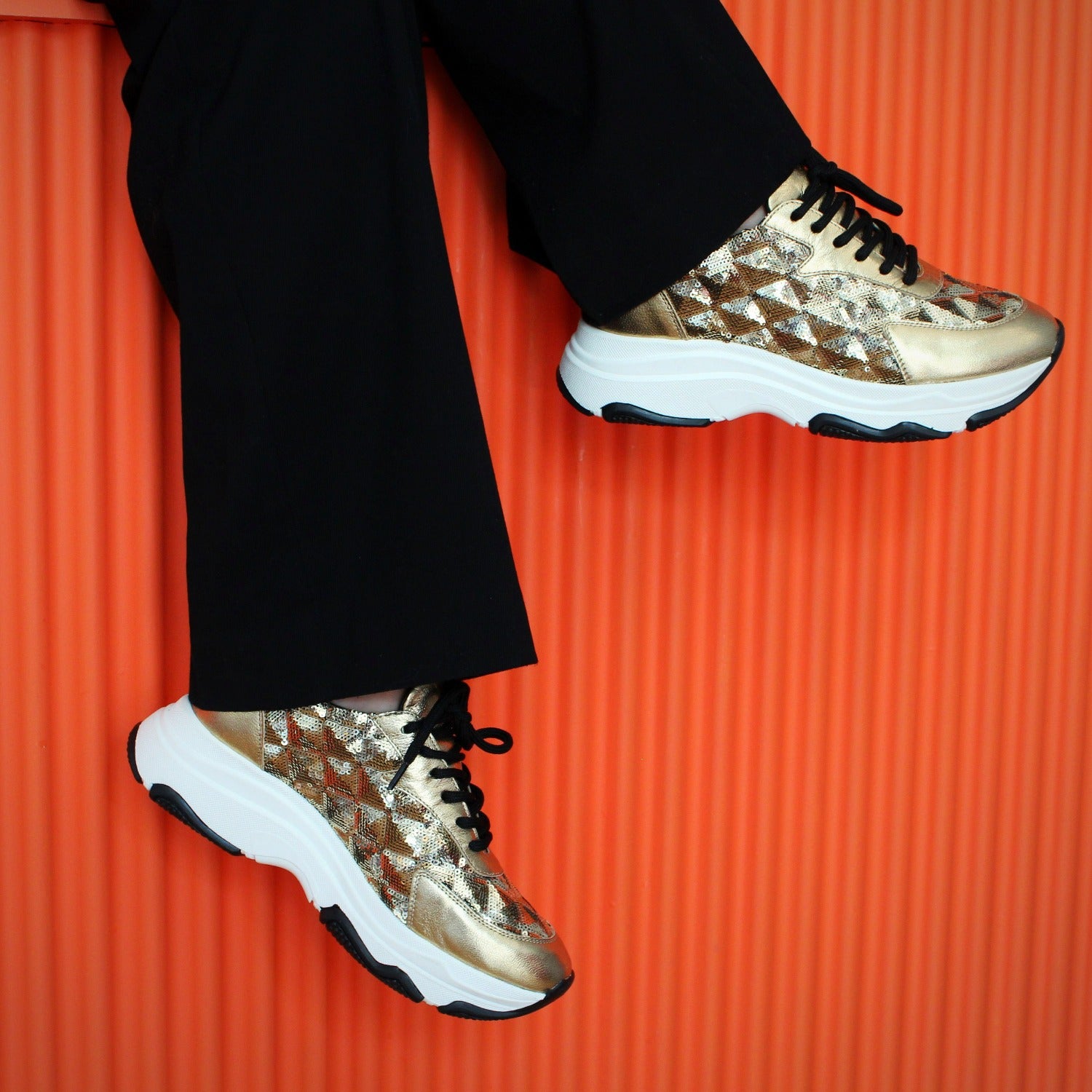 Guld Sneakers med vit sula. Orange vägg. Svarta byxor. The Shoe Bakery